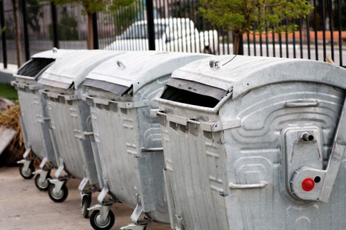 Four large, industrial metal wheelie bins with hinged lids are aligned in a row on a paved surface, likely outside a commercial or residential property in Waltham Forest. The bins are made of galvanized steel with a textured, slightly reflective finish, showing signs of weathering and minor dents. Each bin has black plastic wheels with metal axles for mobility, and the lids are curved and fitted with metal latches. The first bin on the left has its lid partially open, revealing a dark interior. To the right of the bins, a small circular access panel with a red and black knob is visible. In the background, a black metal fence, some greenery, and a parked white van can be seen, indicating a typical urban or suburban environment. The scene appears to be during daylight hours with natural lighting, emphasizing the utilitarian purpose of these waste collection containers, consistent with rubbish removal services offered by companies like Office Clearance Waltham Forest in the local area.