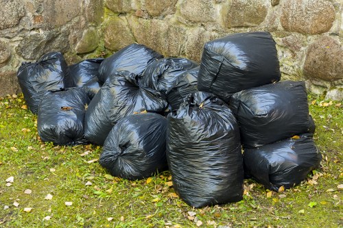 A collection of multiple black plastic rubbish bags, tightly sealed and piled on a grassy outdoor area, with some bags leaning against a rough stone wall. The bags appear full and are made of durable, glossy plastic, reflecting light slightly. The environment shows patches of fallen leaves and moss on the ground, indicating an outdoor setting typical for waste disposal or rubbish removal in Waltham Forest, London. The scene suggests a department or residential area awaiting collection or disposal, managed by a waste removal service such as Office Clearance Waltham Forest. The bags vary in size but are uniformly black, neatly stacked with some overlap, creating a clear focus on waste ready for collection. The background stone wall adds a rustic texture, contrasting with the smooth plastic of the rubbish bags, completing a straightforward depiction of a rubbish collection scene in a local outdoor setting.