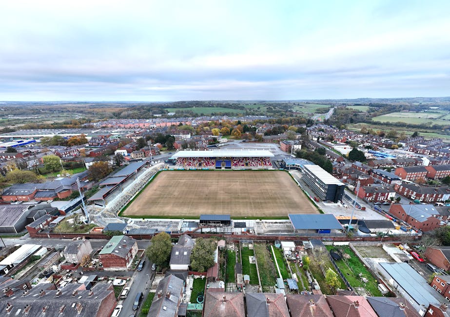 Aerial view of Leyton Orient football stadium in Waltham Forest, showing the rectangular playing field with worn, patchy grass surrounded by a concrete perimeter. The stadium has a seating area with m