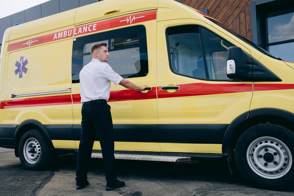 A male paramedic wearing a white uniform shirt stands beside a yellow ambulance with red and black markings, positioned outside a modern building with glass windows and wooden paneling. The paramedic