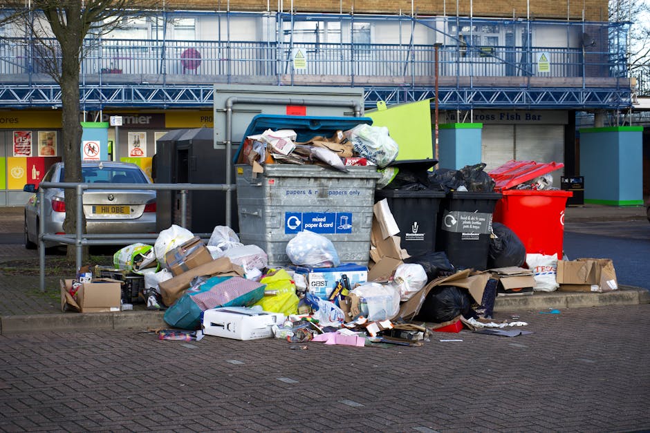 The image depicts a large interior room with white walls and a partially exposed ceiling, filled with a significant accumulation of discarded rubbish. The floor is completely covered with an untidy ar